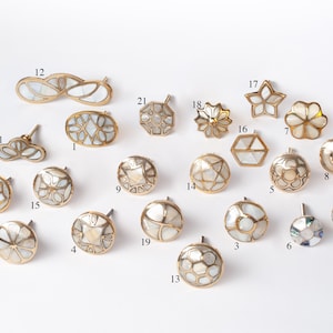 May include: A selection of decorative cabinet knobs and pulls in diverse shapes, including round, oval, and floral designs. The knobs have a gold-coloured metal base with inlaid mother-of-pearl accents. The collection includes a variety of sizes and styles.
