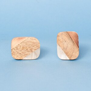 May include: Two square cabinet knobs with rounded corners. Each knob features a combination of wood, copper, and a pearlescent white material. The wood grain is visible, and the knobs are set against a light blue background.