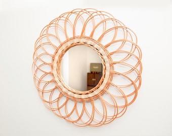 Op de afbeelding: Een ronde wandspiegel met een frame van lichtbruin gevlochten rotan. Het frame bestaat uit meerdere overlappende cirkelvormige lussen die een bloemachtig patroon vormen rondom het centrale ronde glazen spiegeloppervlak.