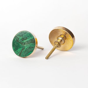 May include: Two cabinet knobs. One has a round, green malachite stone set in a gold-coloured metal frame. The other is gold-coloured metal with a threaded post. The knobs are on a white background. The malachite stone has a swirling pattern.
