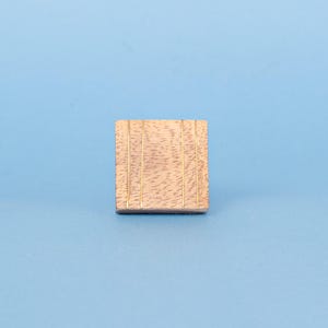 May include: A small, square wooden block with a light brown finish and vertical grooves. The block is set against a solid light blue background, creating a simple, clean aesthetic.