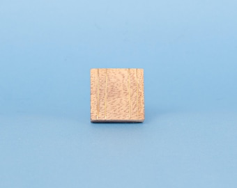 Op de afbeelding: Een klein, vierkant houten blok met een lichtbruine afwerking en verticale groeven. Het blok staat tegen een effen lichtblauwe achtergrond, wat een eenvoudige, strakke esthetiek creëert.