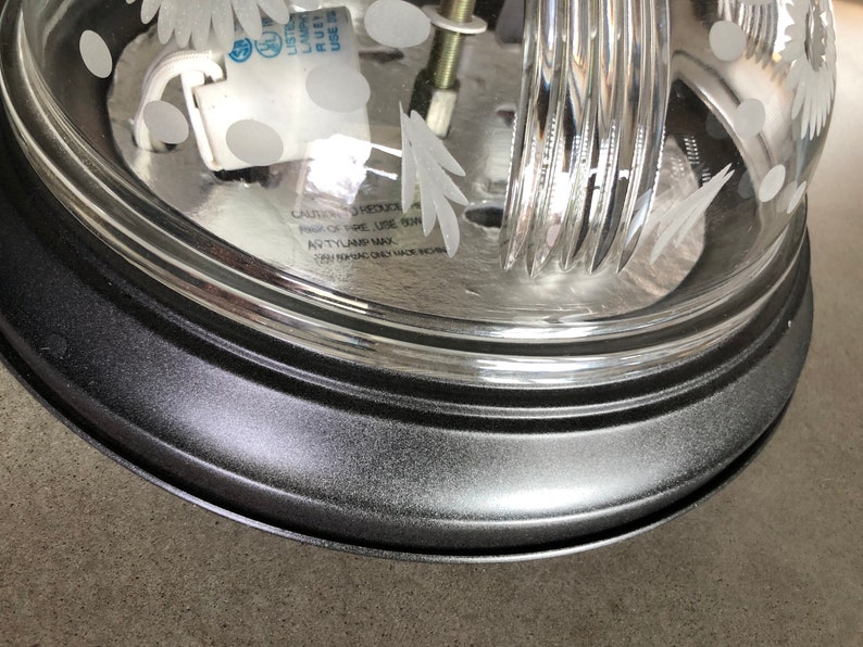 May include: A black metal ceiling light fitting with a clear glass shade featuring a floral design. The shade has a textured surface and a black metal rim. The light fitting has a white electrical socket and a warning label that reads "CAUTION: TO REDUCE RISK OF FIRE, USE ONLY TYPE "A" BULB, MAX 60W."