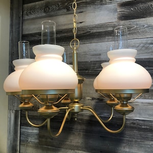 May include: A vintage-style chandelier with five lights. The fixture has a gold-colored metal frame and arms, with frosted glass shades and clear glass chimneys. The chandelier is suspended from a gold chain.