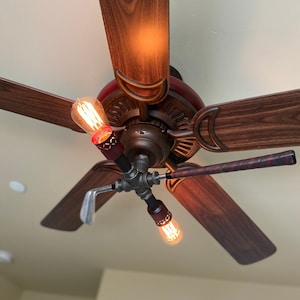 May include: A ceiling fan with wooden blades and a brown metal base. The fan has Edison bulb light fixtures with golf club arms.