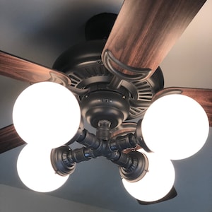 May include: A black ceiling fan with four white globe light bulbs. The fan blades are made of wood and have a dark brown finish. The light fixture is made of black metal pipes.