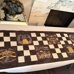May include: A rectangular, dark brown and cream checkered table runner. The runner features floral and bee-themed designs, with text that reads "Honey Bees and Flowers please" and "Bee Kind".