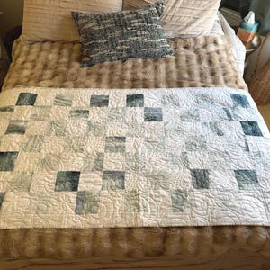 May include: A handmade quilt with a patchwork design. The quilt features squares of varying shades of blue and green on a white background. The quilt is placed on a bed with a faux fur blanket and decorative pillows.