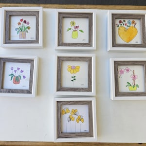 May include: Seven framed watercolor paintings of flowers. Each painting features a different floral arrangement in a vase or container. The frames are white with a brown inner border. The paintings are on a white surface.