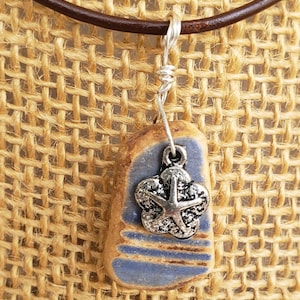 May include: A blue and brown ceramic pendant with a silver starfish charm hangs from a brown leather cord. The pendant is a unique and handmade piece of jewelry.