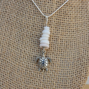 May include: A silver turtle pendant necklace with a stack of white shell beads. The necklace is displayed on a brown burlap background.