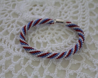 May include: A red, white, and blue beaded bracelet with a silver clasp. The bracelet is made with small, round beads and has a spiral pattern.