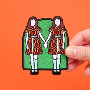 Könnte beinhalten: Ein gestickter Aufnäher mit zwei Mädchen in roten und orangen Kleidern mit einem geometrischen Muster. Die Mädchen halten sich an den Händen und stehen vor einem grünen Hintergrund.