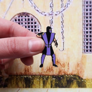 May include: A black and purple enamel pin of a video game character in a fighting pose. The character is wearing a purple and black outfit with a black mask.