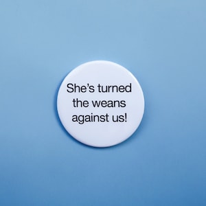 May include: White button with black text that reads "She's turned the weans against us!"