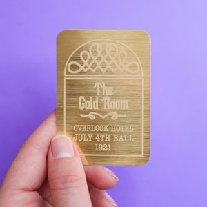 Puede incluir: Una tarjeta de metal de color dorado con el texto "The Gold Room" y "Overlook Hotel July 4th Ball 1921" grabado en ella.
