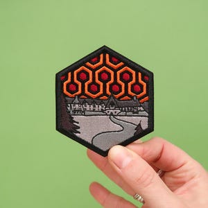 May include: A hexagonal embroidered patch depicting a stylised image of the Overlook Hotel from the film "The Shining". The hotel is shown in the background with a winding road leading up to it. The foreground features a red and orange geometric pattern.