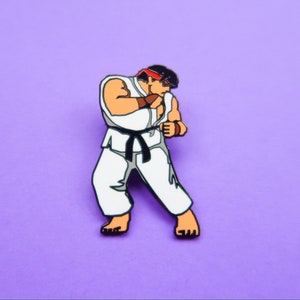May include: An enamel pin featuring a cartoon character in a white karate uniform with a black belt, red headband, and brown hair. The character is in a fighting pose with their fist raised.