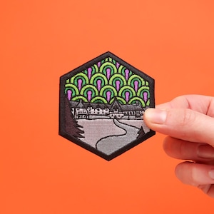 Puede incluir: Parche bordado en blanco y negro con un diseño verde y morado. El parche presenta un edificio con un camino sinuoso que conduce hasta él. El diseño recuerda al Hotel Overlook de la película "El Resplandor".