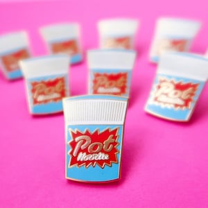 May include: A close-up of a gold-coloured enamel pin shaped like a cup of instant noodles. The pin is blue and white with a red and gold "Pot Noodle" logo. The pin is on a pink background.