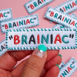 May include: A white fabric patch with green and white striped trim. The patch has the text "BRAINIAC" in red, black, and green letters with quotation marks around the text.