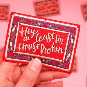 May include: Red embroidered patch with a white border and purple and gold accents. The patch says "Hey at least I'm house broken".