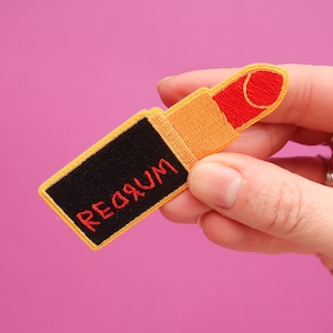 May include: A red and yellow embroidered patch shaped like a lipstick tube with the word "REDRUM" written in red on the black case.