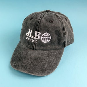 Könnte beinhalten: Eine schwarze, abgenutzte Baseballkappe aus Baumwolle mit einem weißen gestickten Logo, auf dem "JLB CREDIT" und eine Weltkugel abgebildet sind.