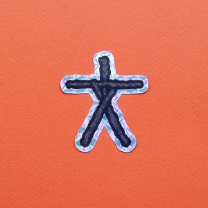 May include: A sticker with a black and silver design resembling a person with outstretched arms. The sticker has a silver border and is set against an orange background. The design is a stylized representation.