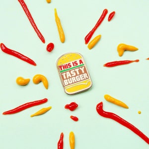 May include: A gold-colored enamel pin with a cartoon burger and the text "This is a Tasty Burger". The pin is surrounded by red and yellow paint splatters on a light blue background.
