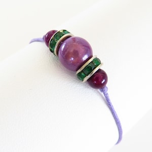 May include: A purple and green beaded bracelet with a large purple bead accented with green rhinestones. The bracelet is made with a lavender cord.