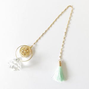 Op de afbeelding: Een goudkleurige ketting met een decoratieve hanger met een helder kristal, een goudkleurige muntachtige bedel en een mintgroene kwast. De hanger heeft een cirkelvormig ontwerp met een kleinere muntachtige bedel binnenin.