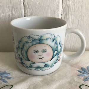 Peut inclure: Une tasse en céramique blanche avec un motif de poupée de chou bleu et vert. La tasse présente un visage de bébé souriant avec des yeux bleus et un fard à joues rose.