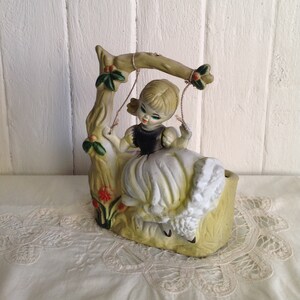 May include: A ceramic figurine of a girl with blonde hair and a white dress sitting on a swing. The swing is attached to a tree branch with green leaves. The figurine is painted in shades of white, green, and brown.