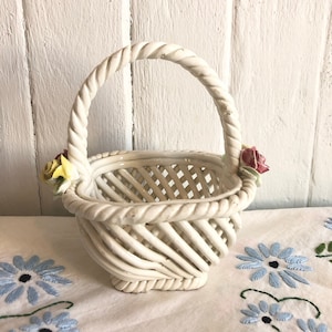 Peut inclure: Un panier en céramique blanche avec un motif tissé et une poignée en corde torsadée. Le panier a deux petites décorations florales sur les côtés.