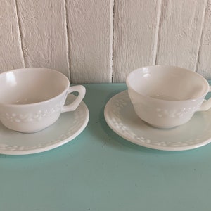 Puede incluir: Dos tazas de té y platillos de vidrio de leche blanco con un diseño floral. Las tazas y los platillos están sobre una superficie azul claro.