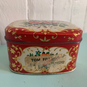 May include: Red and white tin box with a floral design and gold accents. The box has a label that reads "Tom Fill" and "Ed Toffees Assorted".