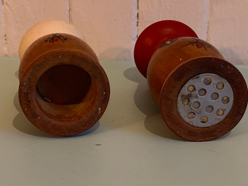 May include: Two wooden salt and pepper shakers with a red and white top and a metal grinder on the bottom.