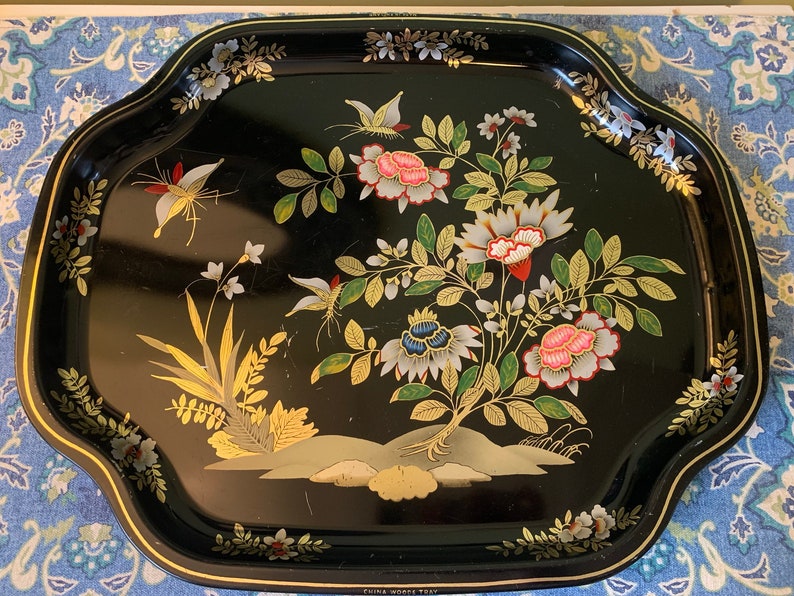 Pu&ograve; includere: Vassoio da portata in metallo nero con bordo dorato e un motivo floreale con farfalle, fiori e foglie in tonalit&agrave; di rosa, bianco, blu e verde. Il vassoio ha le parole "Fine Wooden Tray" stampate sul fondo.