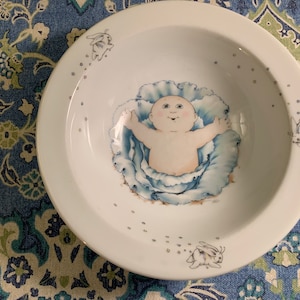 Può includere: Una ciotola in ceramica bianca con un'illustrazione blu e bianca di un bambino in un cavolo. La ciotola ha un bordo bianco ed è decorata con piccoli punti bianchi intorno al bordo.