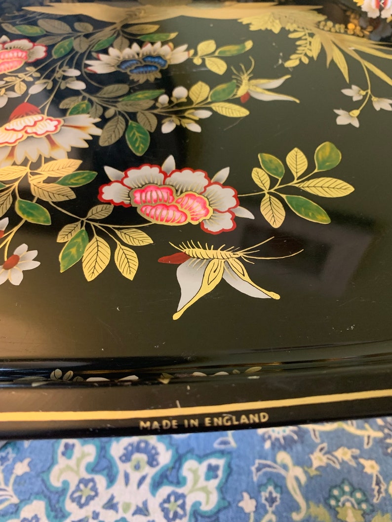 Pu&ograve; includere: Vassoio in metallo nero con bordo dorato e design floreale. Il vassoio presenta una farfalla bianca e le parole "Made in England" sul fondo.