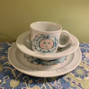 Può includere: Un set di piatti in ceramica bianca per bambini con un design floreale blu e bianco. La tazza presenta un viso di bambino dei cartoni animati con un cappello floreale blu e bianco.