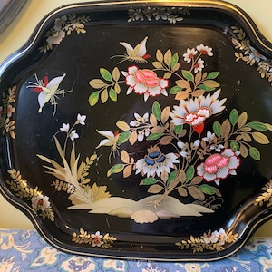 Pu&ograve; includere: Un vassoio da portata in metallo nero con un bordo dorato e un motivo floreale con farfalle, fiori e foglie in tonalit&agrave; di rosa, bianco, verde e oro.