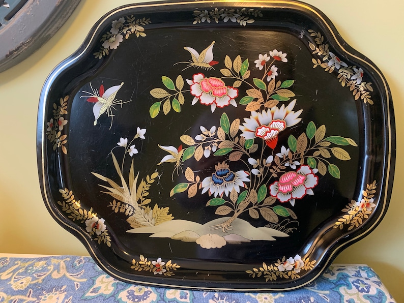 Pu&ograve; includere: Un vassoio da portata in metallo nero con un bordo dorato e un motivo floreale con farfalle, fiori e foglie in tonalit&agrave; di rosa, bianco, verde e oro.