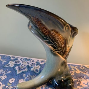 Peut inclure: Un vase en céramique bleu et vert en forme de tête d'oiseau avec un long bec. Le vase est décoré de pommes de pin et d'aiguilles brunes et jaunes.
