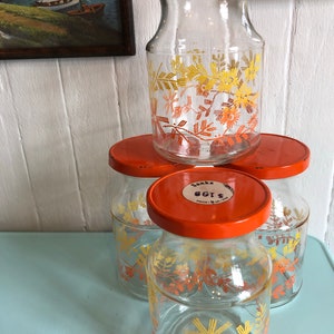 Puede incluir: Conjunto de cuatro tarros de vidrio con tapas naranjas y dise&ntilde;os florales. Los tarros son de vidrio transparente con un patr&oacute;n floral amarillo y naranja. Las tapas son de color naranja brillante. Los tarros est&aacute;n etiquetados con "Sanka Eggs" en la tapa.