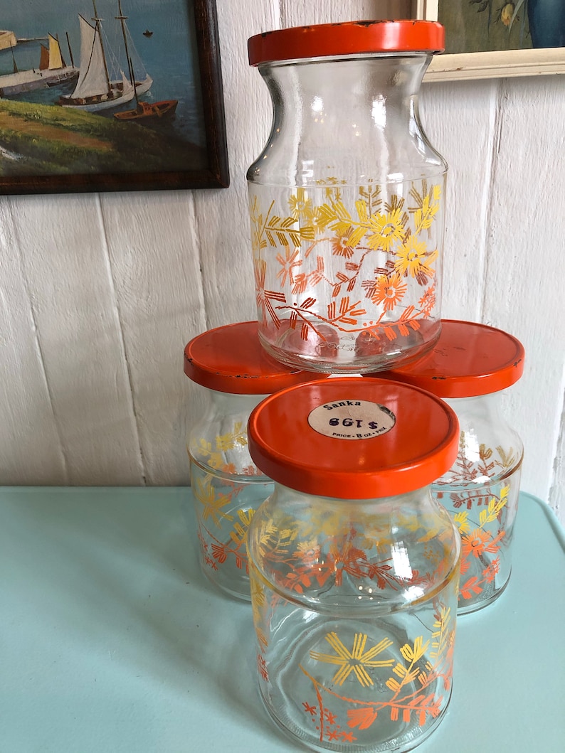 Puede incluir: Conjunto de cuatro tarros de vidrio con tapas naranjas y dise&ntilde;os florales. Los tarros son de vidrio transparente con un patr&oacute;n floral amarillo y naranja. Las tapas son de color naranja brillante. Los tarros est&aacute;n etiquetados con "Sanka Eggs" en la tapa.