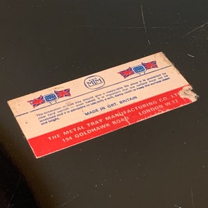 Pu&ograve; includere: Un'etichetta rosso, bianco e blu con il testo "The Metal Tray Manufacturing Co. Ltd. 194 Goldhawk Road London W.12" e "Made in Grt. Britain". L'etichetta presenta anche un logo con le lettere "MM" e le parole "Elite Trays".