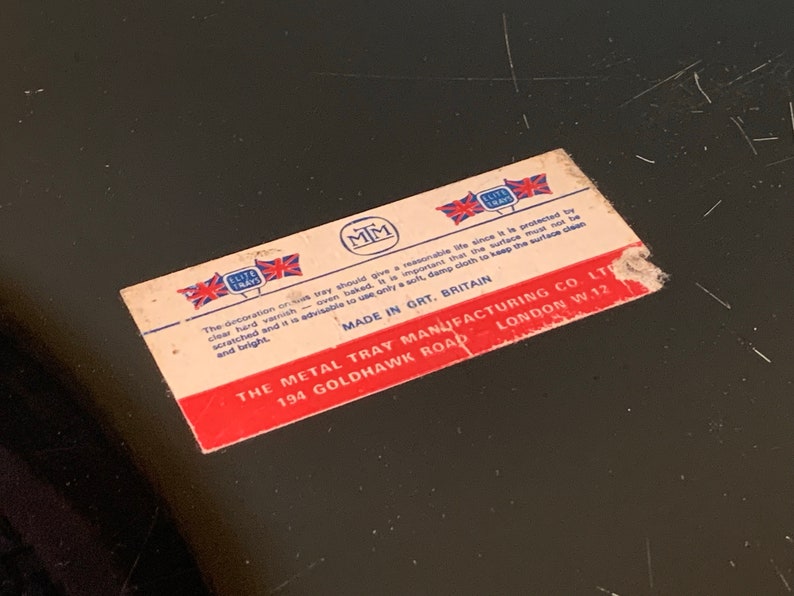 Pu&ograve; includere: Un'etichetta rosso, bianco e blu con il testo "The Metal Tray Manufacturing Co. Ltd. 194 Goldhawk Road London W.12" e "Made in Grt. Britain". L'etichetta presenta anche un logo con le lettere "MM" e le parole "Elite Trays".