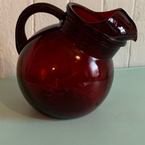 Peut inclure: Un pichet en verre rouge avec un corps rond et un bec large. Le pichet a une poignée et un couvercle.